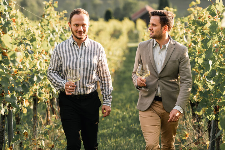 Zwei Männer stehen zwischen Weinreben bei Sonnenschein. Sie halten jeweils ein Weinglas in den Händen.