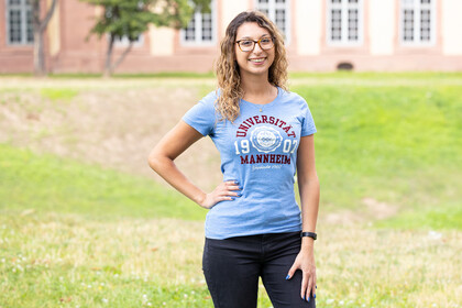 Eine lächelnde Person trägt ein hellblaues T-Shirt mit dem Logo der Uni Mannheim und steht auf der Mensawiese vor dem Schloss. Die Person heißt Anais Bentayeb..