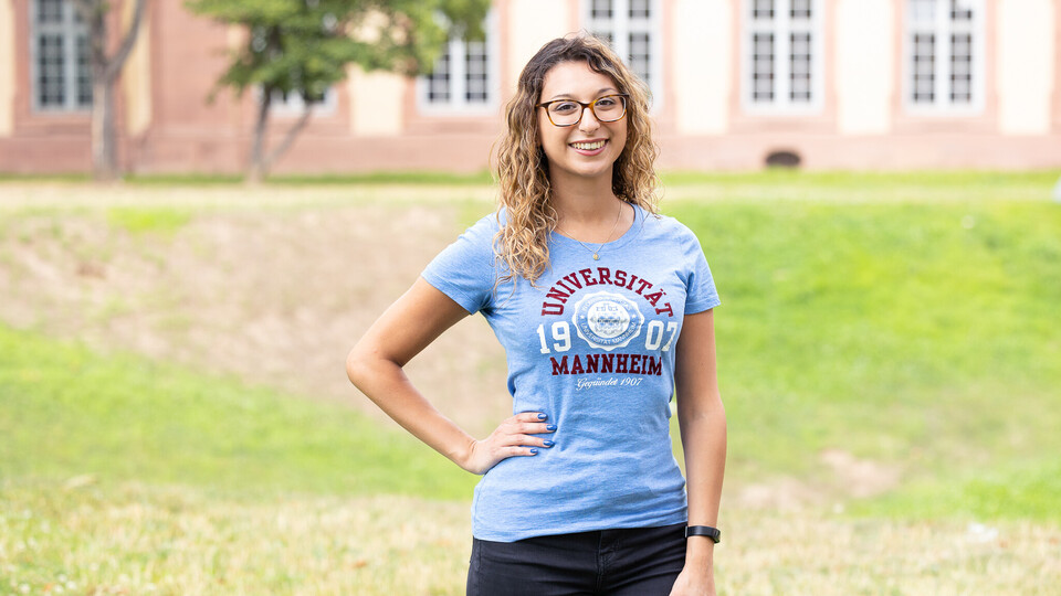 Eine lächelnde Person trägt ein hellblaues T-Shirt mit dem Logo der Uni Mannheim und steht auf der Mensawiese vor dem Schloss. Die Person heißt Anais Bentayeb..