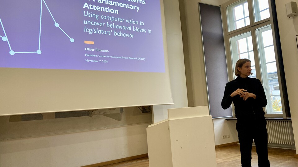 Oliver Rittmann gives his lecture. He is wearing a black sweater and black trousers. You can see his presentation on the screen in the background.