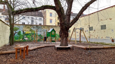 Eine ruhige Spielecke im Freien mit einem großen, blattlosen Baum in der Mitte, umgeben von Spielgeräten wie Rutschen und Schaukeln. Die Wand im Hintergrund ist mit einem Wandgemälde von Pflanzen und Tieren bemalt.