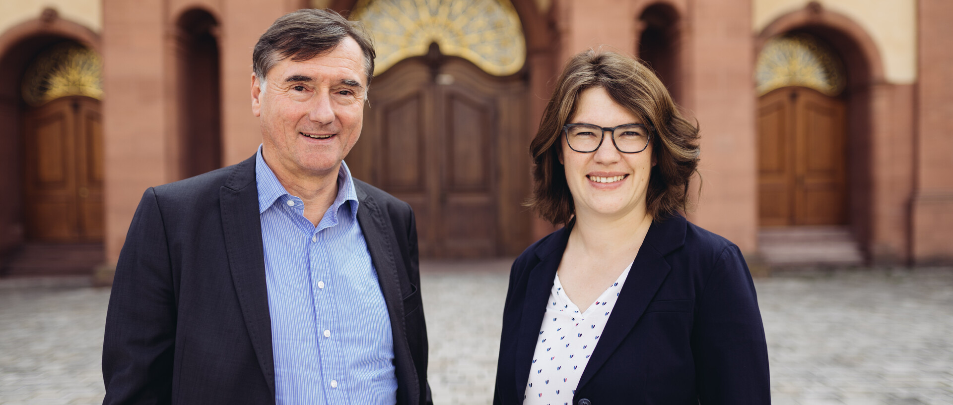 Portraitfoto von zwei Personen. Im Hintergrund das Schloss