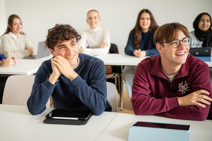 Sieben Studierende in einem Seminarraum. Sie sitzen an Tischen udn hören jemandem zu. Sie lachen und sind positiv gestimmt.