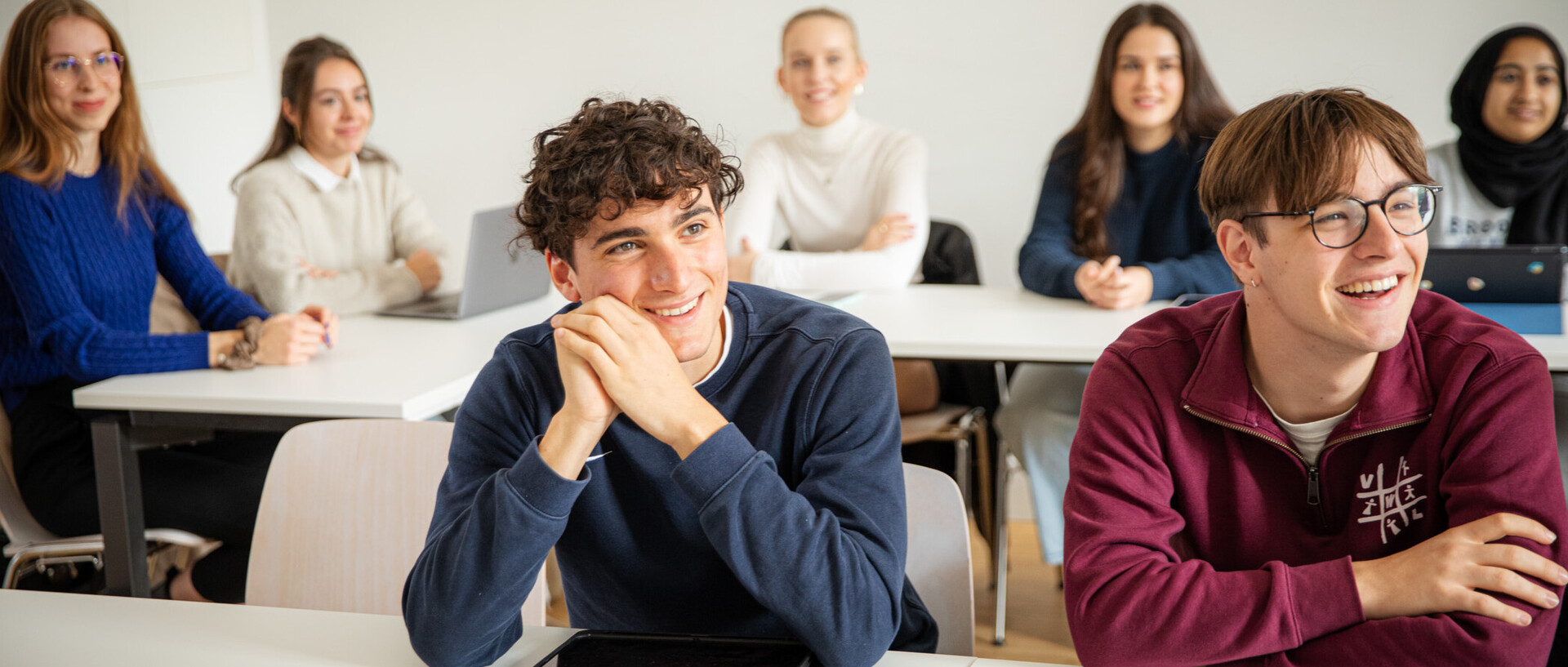 Sieben Studierende in einem Seminarraum. Sie sitzen an Tischen udn hören jemandem zu. Sie lachen und sind positiv gestimmt.