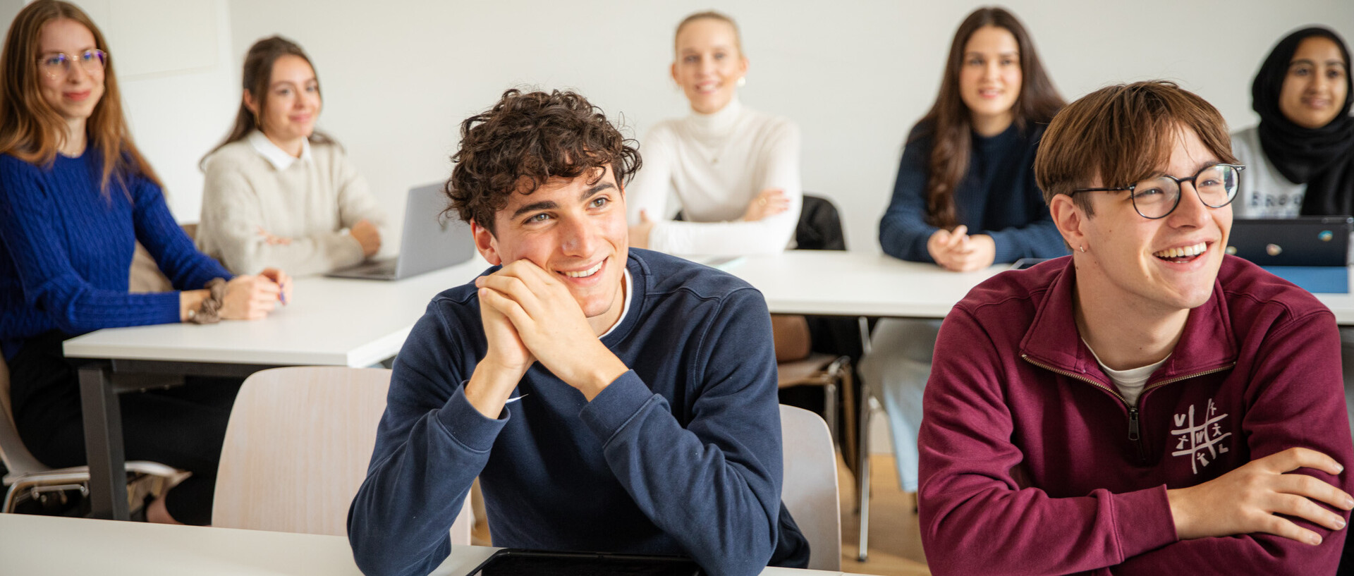 Sieben Studierende in einem Seminarraum. Sie sitzen an Tischen udn hören jemandem zu. Sie lachen und sind positiv gestimmt.
