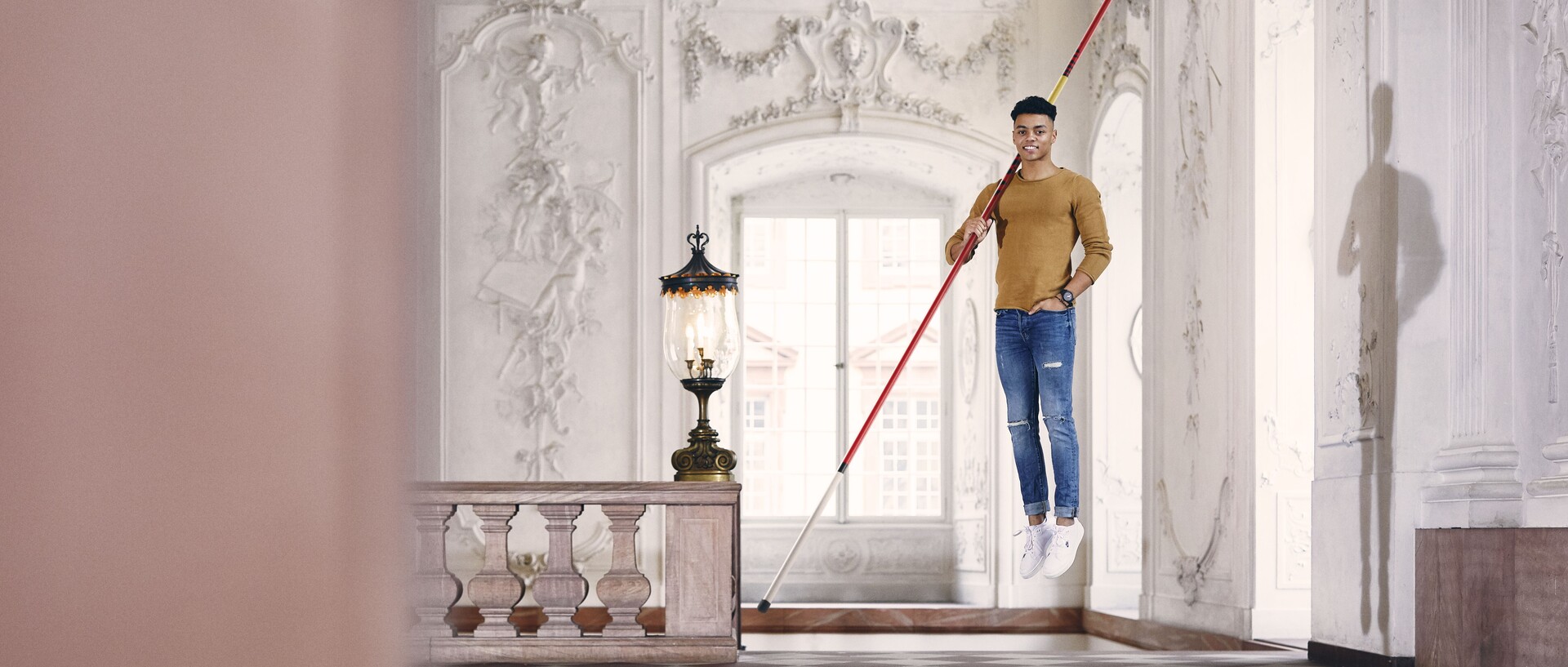 Ein Mann mit einem langen Stab über der Schulter schwebt über dem Boden. Im Hintergrund ein Treppengeländer, große Fenster und ornamentierte Wände.