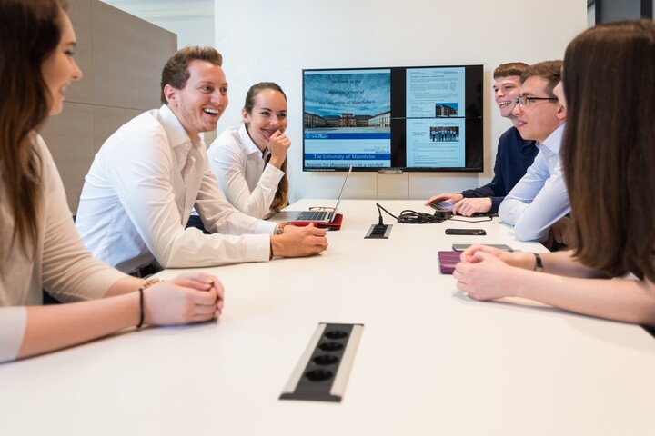 6 Personen, die mit Laptops an einem Tisch sitzen und miteinander sprechen. Sie tragen weiße Hemden bzw. Blusen. 