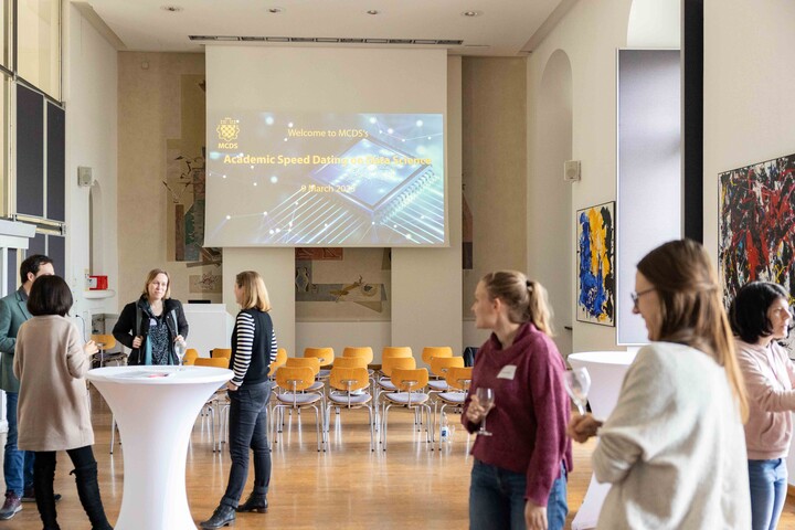 Festsaal der Universität Mannheim mit mehreren Stuhlreihen. Davor stehen Personen, die sich unterhalten. Im Hintergrund ist eine Leinwand, auf der man das Logo des MCDS sieht sowie den Titel der Veranstaltung.