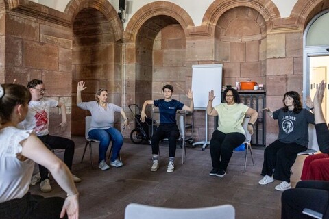 Personen im Stuhlkreis in den Katakomben der Universität. Alle heben beide Arme an, während bei einem der beiden die Hand nach oben zeigt und die andere nach unten.