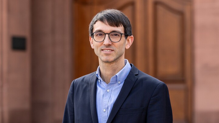 Sebastian Stier steht vor einer dunklen Holztür. Er trägt einen dunkelblauen Sakko auf hellblauem Hemd und lächelt in die Kamera.