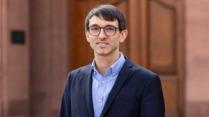 Sebastian Stier steht vor einer dunklen Holztür. Er trägt einen dunkelblauen Sakko auf hellblauem Hemd und lächelt in die Kamera.