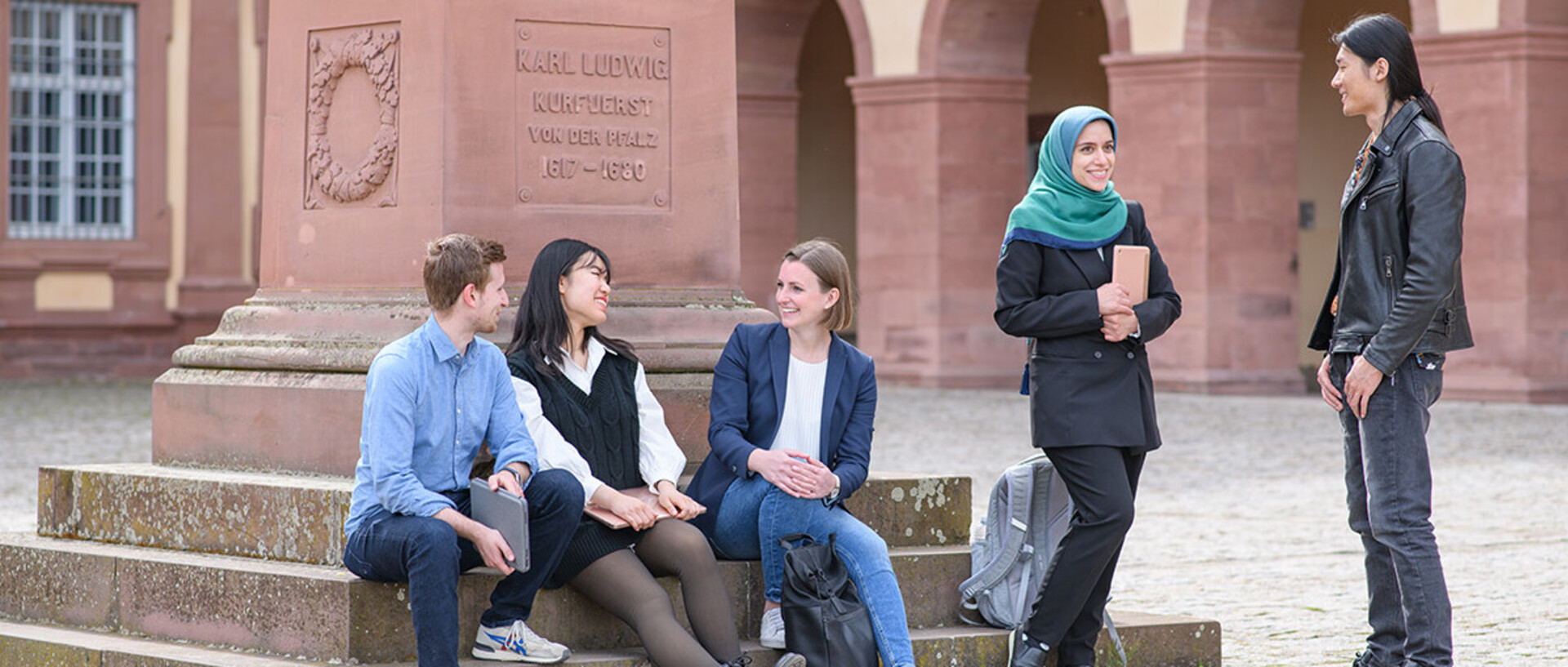 Five students conversing by the statue of Prince-Elector Karl Ludwig on the Ehrenhof of the Palace. Three are sitting on the steps of the monument. Two are standing.
