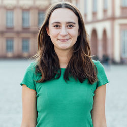 Flavia Saxler hat dunkelbrauenes leicht über die Schulter ragendes Haar. Sie trägt ein grünes T-Shirt und lächelt.