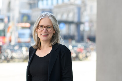 Frau Mata trägt ein schwarzes Oberteil mit einem schwarzen Sakko und eine Brille.
