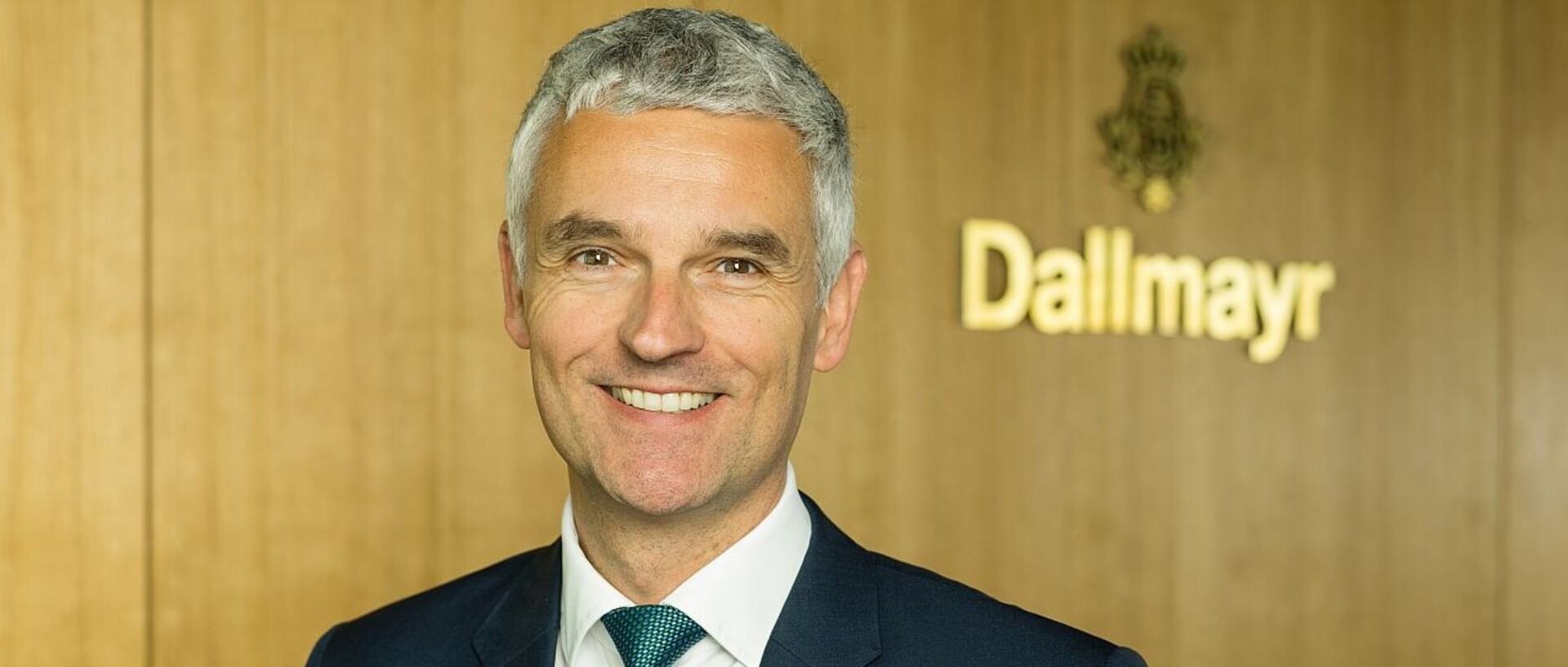 Dr. Johannes Dengler lächelnd in schwarzem Anzugf, weißem Hemd und grüner Krawatte vor einer Holzwand mit dem Schriftzug "Dallmayr"