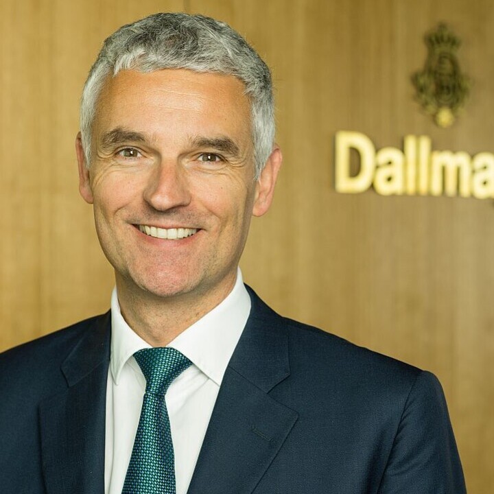Dr. Johannes Dengler lächelnd in schwarzem Anzugf, weißem Hemd und grüner Krawatte vor einer Holzwand mit dem Schriftzug "Dallmayr"