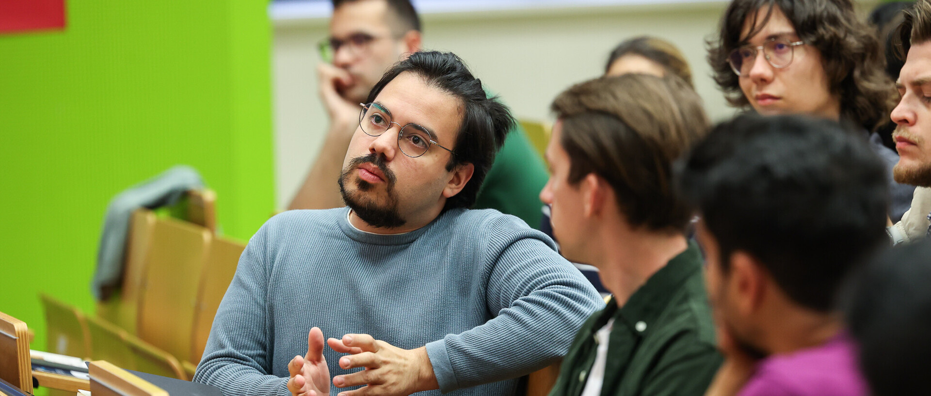 Studierende sitzen in einem Hörsaal. Ein Student mit Bart spricht und gestikuliert mit seinen Händen.