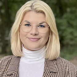 Die Bachelorabsolventin Jule Schabernack trägt kinnlanges blondes Haar, eine dezente, runde Brille und einen karierten Blazer über einem weißen Rollkragenpullover.