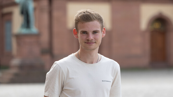 Jan-Niklas Otte. He is wearing a white T-shirt and is standing on the Ehrenhof in front of the castle.
