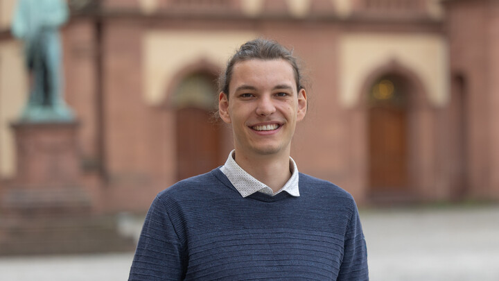 Konrad Strehl. He is wearing a blue sweater over a white shirt and is standing on the Ehrenhof in front of the castle.