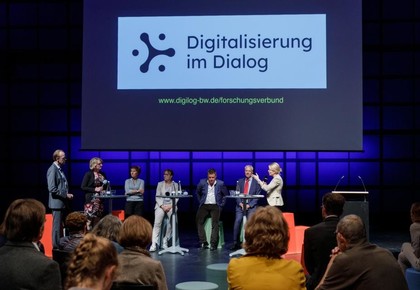 Das Bild zeigt Teilnehmerinnen und Teilnehmer einer Podiumsdiskussion. Im Hintergrund ist auf eine Leinwand projiziert Logo und Schiftzug von Digitalisierung im Dialog.