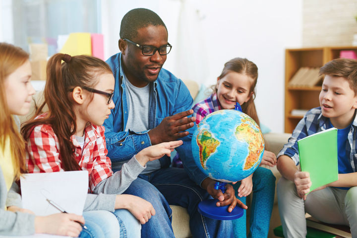 Ein Mann zeigt Hält einen Globus in seiner Hand und zeigt auf ein Land. Vier Kinder schauen ihm interessiert dabei zu.
