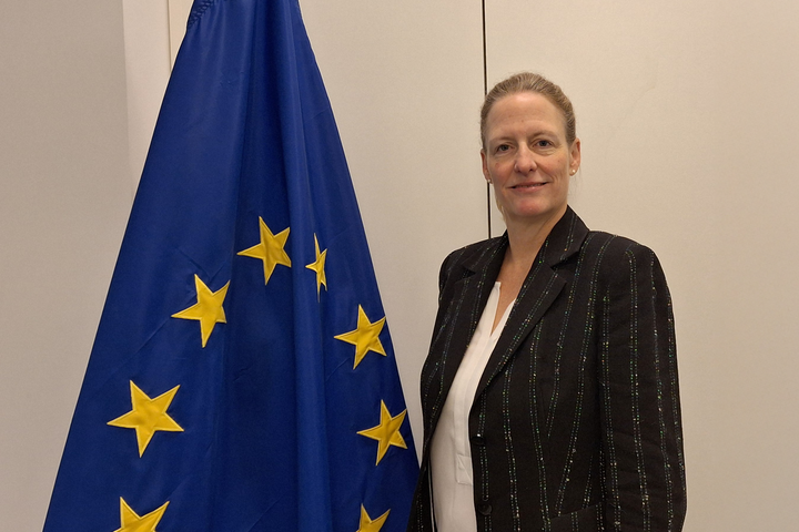Alumna Dr. Alexandra Kiel trägt einen schwarz-gestreiften Blazer und ihre blonden Haare zurückgebunden. Sie steht neben der Europaflagge und lächelt freundlich.