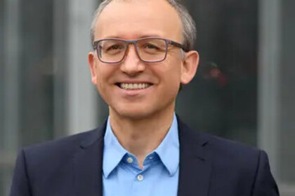 Eine lächelnde Person trägt ein hellblaues Hemd sowie einen dunkelblauen Blazer und steht vor einem Gebäude. Die Person heißt Thomas Gschwend.