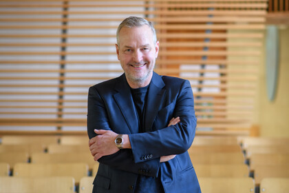 Portrait von Hans Peter Grüner. Er trägt ein dunkelblaues Hemd sowie einen dunkelblauen Blazer und steht mit verschränten Armen in einem leeren Hörsaal.