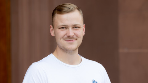 Henning Schatz. He is wearing a white T-shirt and is standing in front of brown wall.