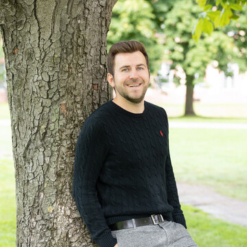 Tim Neubauer. Er lehnt locker an einem Baum und lacht, dabei trägt er einen schwarzen Pullover.