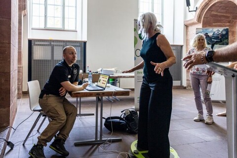 Frau steht im Schloss der Universität auf einer Gleichgewichts-Platte und versucht das Gleichgewicht zu halten. Daneben sitzt Mann auf einem Stuhl, bedient die Gleichgewichts-Platte per Laptop und schaut die Frau an.