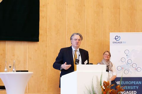 Eine Person trägt einen schwarzen Blazer und spricht hinter einem Rednerpult. Hinter ihm steht ein Banner von Engage.Eu.