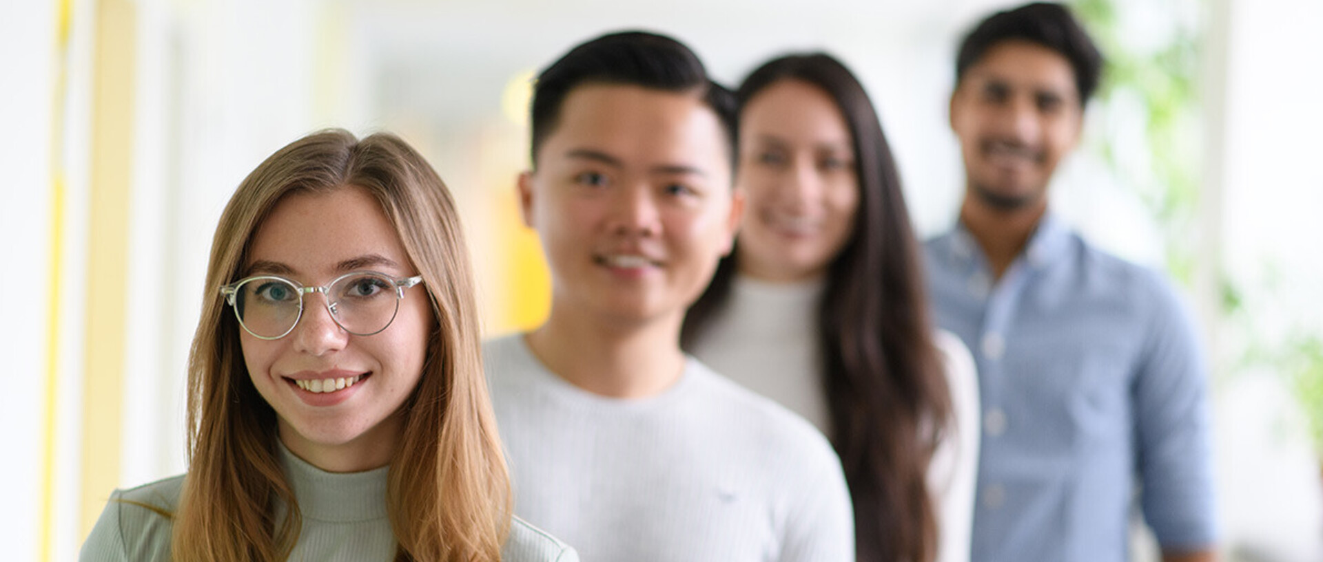 Zwei Studentinnen und zwei Studenten stehen schräg hintereinander. Nur die vordere Studentin ist scharfgestellt. Sie trägt lange blonde Haare, eine silberne Brille und einen mintfarbenen Rollkragenpullover.