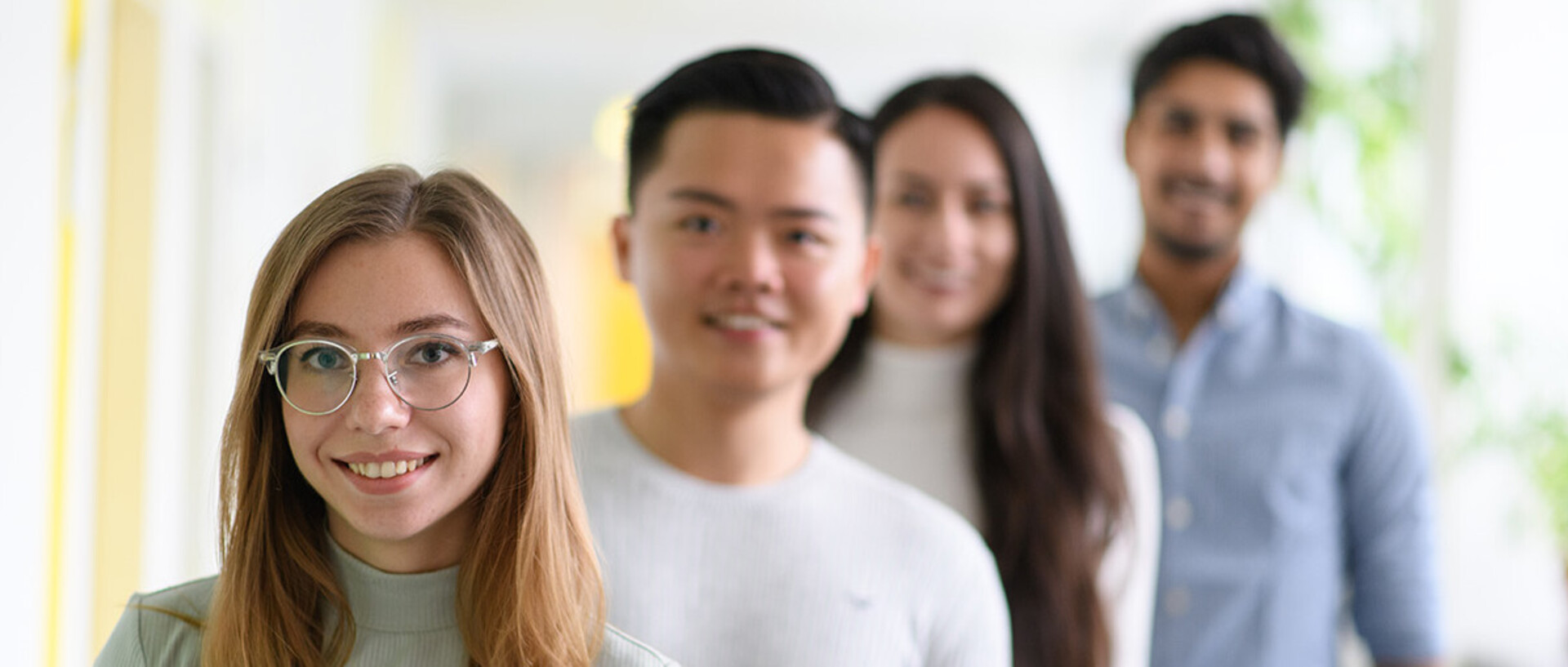 Zwei Studentinnen und zwei Studenten stehen schräg hintereinander. Nur die vordere Studentin ist scharfgestellt. Sie trägt lange blonde Haare, eine silberne Brille und einen mintfarbenen Rollkragenpullover.