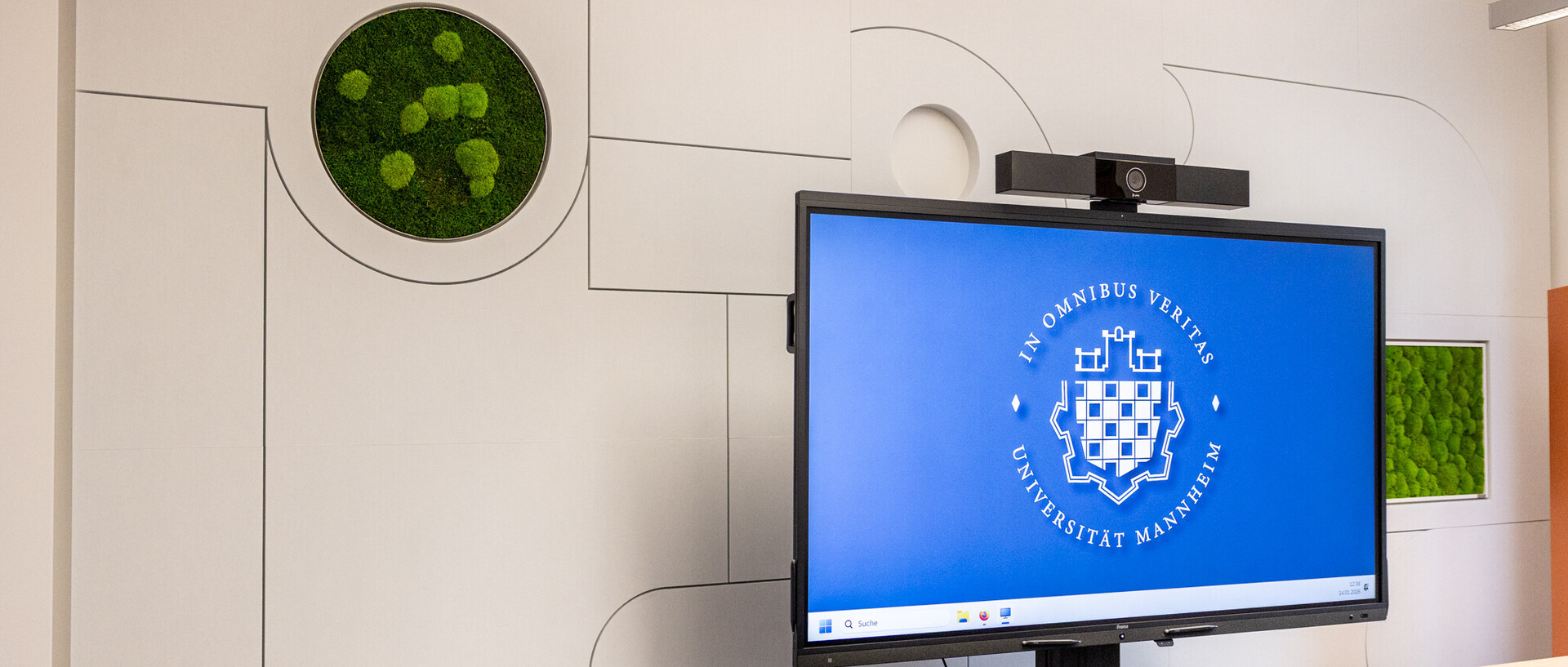 Man sieht einen helle, schallhemmende Wand mit Mooseinsatz. Davor ein großes Display, auf dem das Logo der Uni Mannheim zu sehen ist.