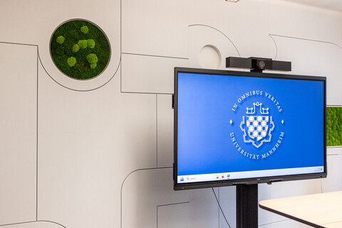 Das Bild zeigt eine schallschluckende Wand mit eingebauten grünen Moos-Elementen. Davor steht ein großes Display mit dem Logo der Uni Mannheim.