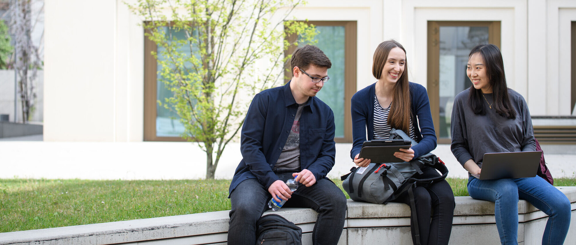 Drei Studierende sitzen lachend auf der Mauer vor dem Gebäude B6 der Universität Mannheim