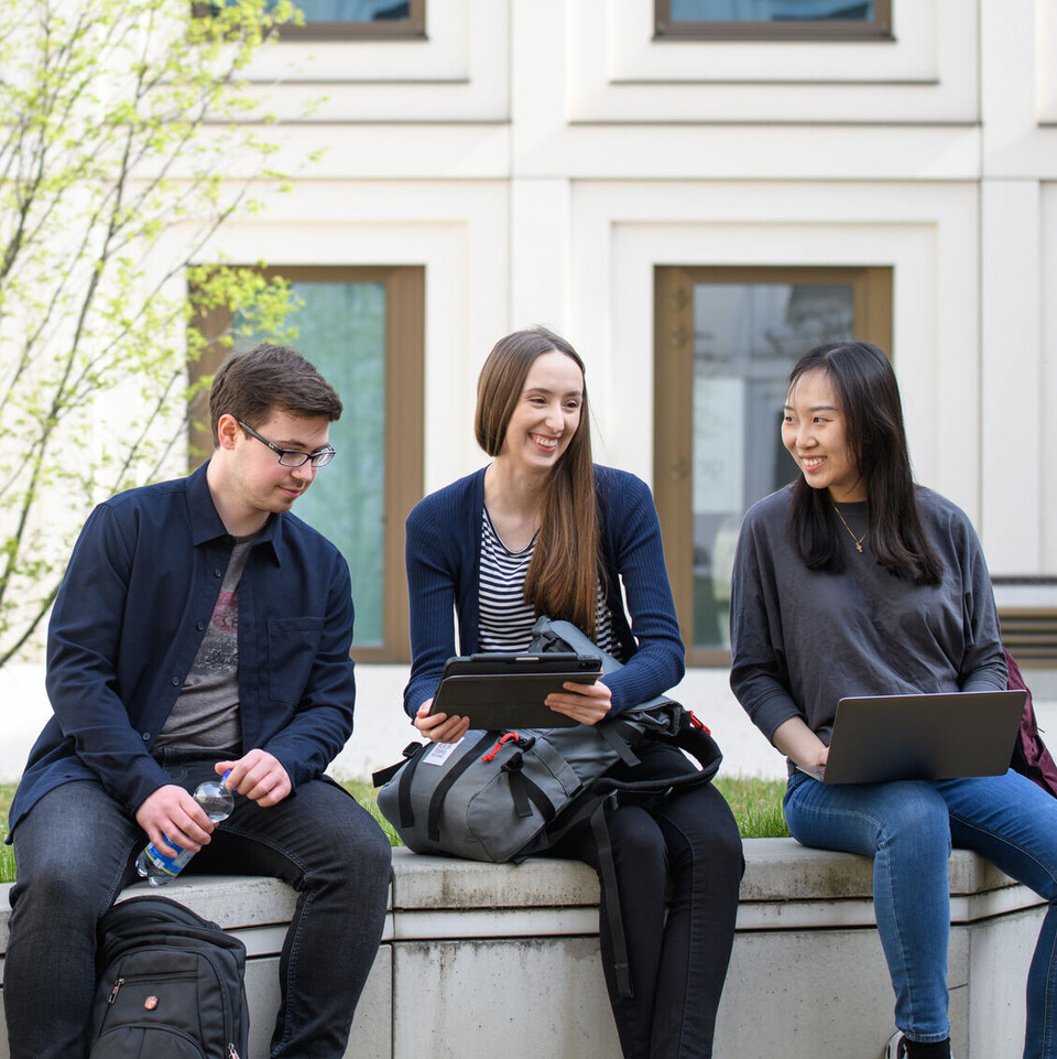 Drei Studierende sitzen lachend auf der Mauer vor dem Gebäude B6 der Universität Mannheim