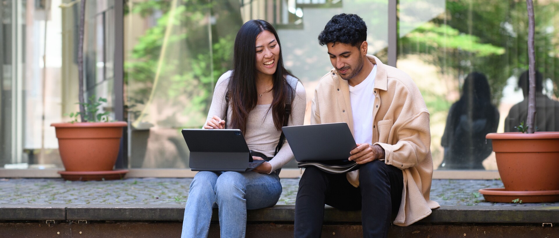 Ein Student und eine Studentin sitzen draußen auf einer Mauer und lernen gemeinsam an ihren Laptops.