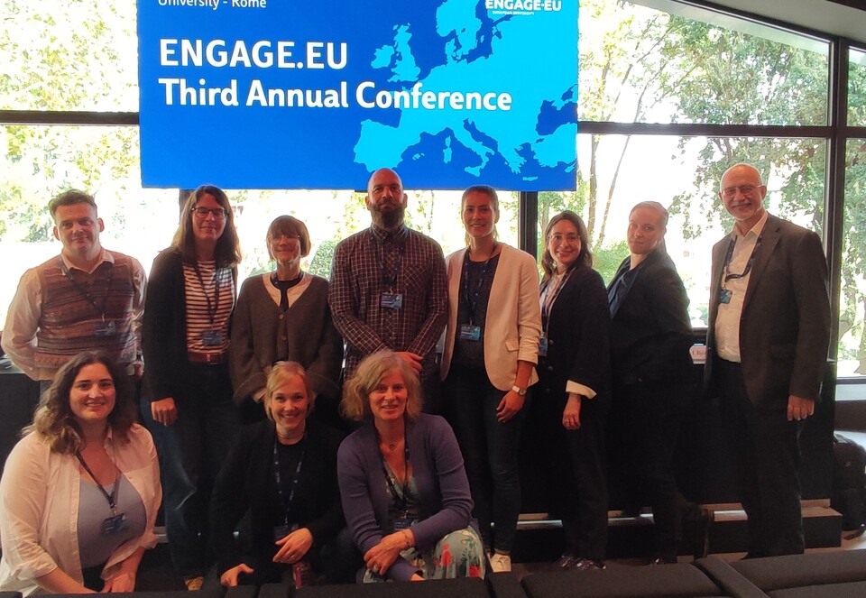 Eine Gruppe von Personen steht nebeneinander vor einem Fenster. Hinter ihnen hängt ein Banenr mit der Aufschrift "Engage.Eu - Third Annual Conference".