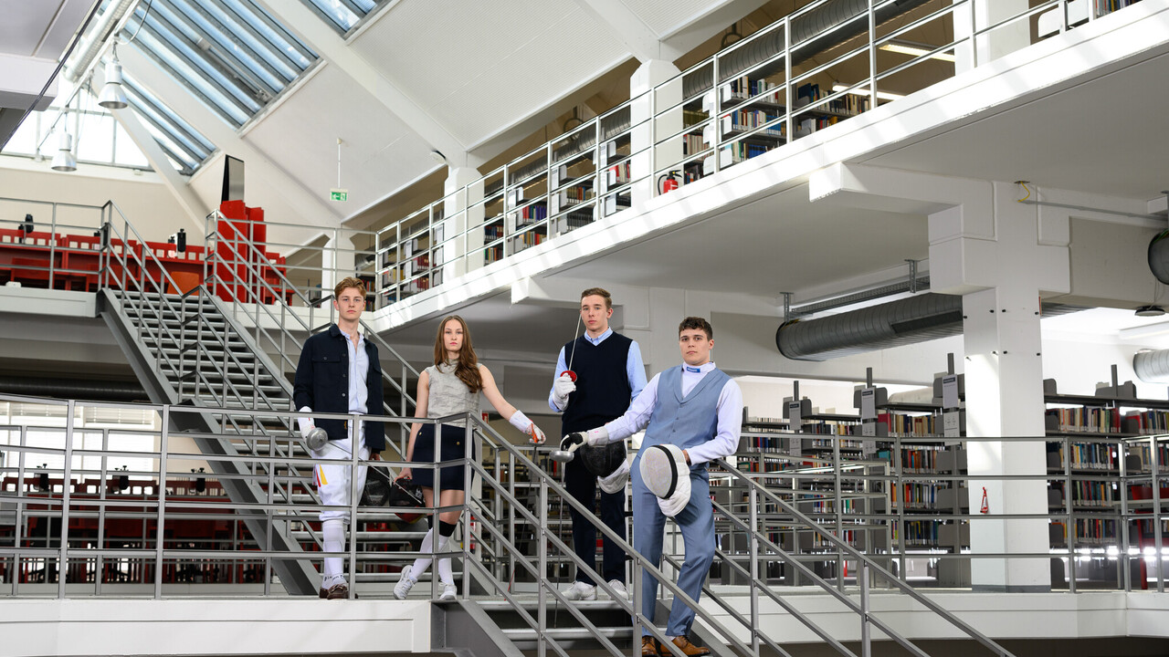 Drei Sportler und eine Sportlerin in Fecht-Ausrüstung und formeller Kleidung stehen am Treppengeländer der A3-Bibliothek.