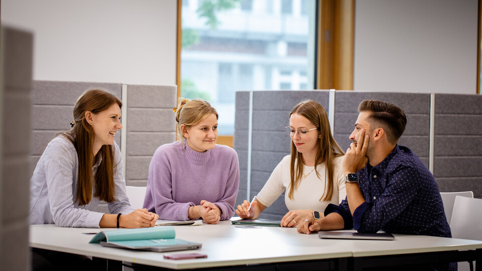 Vier junge Erwachsene sitzen in einem modernen Büro an einem Tisch und diskutieren konzentriert miteinander, vor ihnen liegen Notizbücher und Tablets.