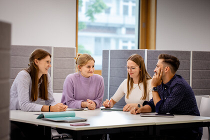 Vier junge Erwachsene sitzen in einem modernen Büro an einem Tisch und diskutieren konzentriert miteinander, vor ihnen liegen Notizbücher und Tablets.