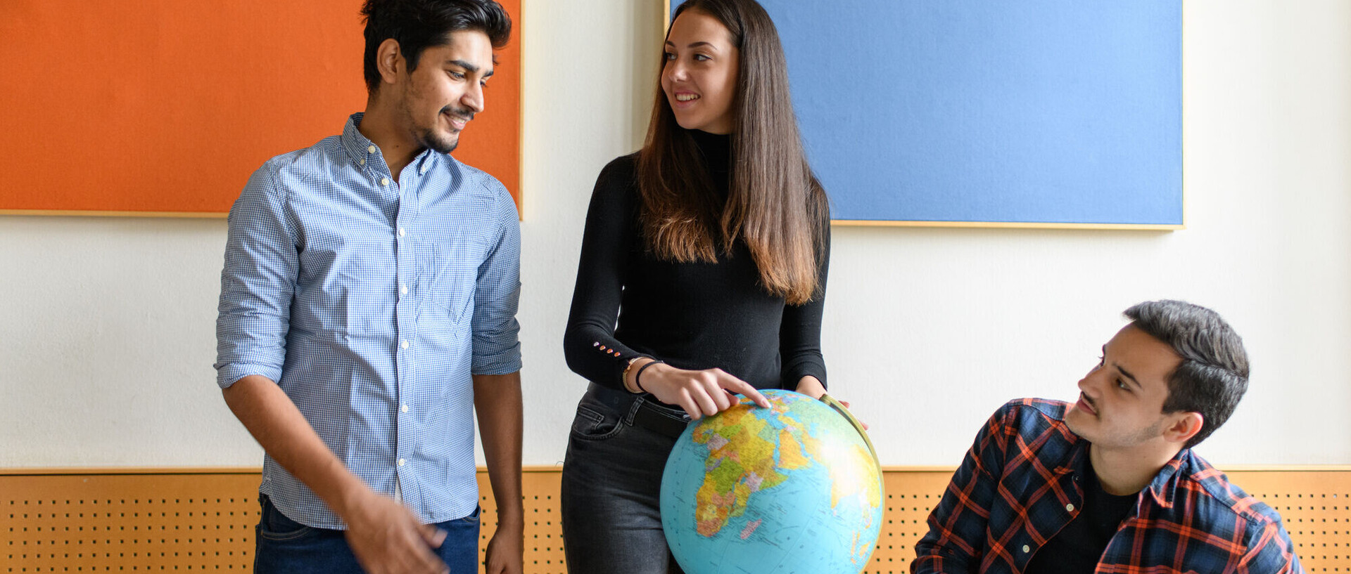 Drei Studierende um eine Weltkugel. Eine Studentin zeigt auf einen Punkt der Weltkugel.