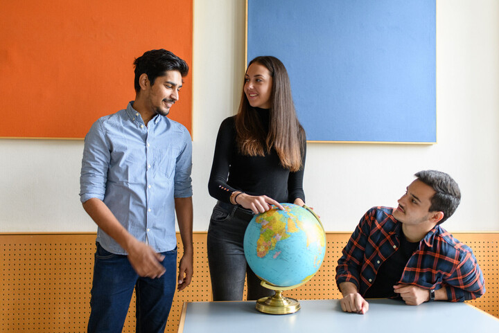 Drei Studierende um eine Weltkugel. Eine Studentin zeigt auf einen Punkt der Weltkugel.