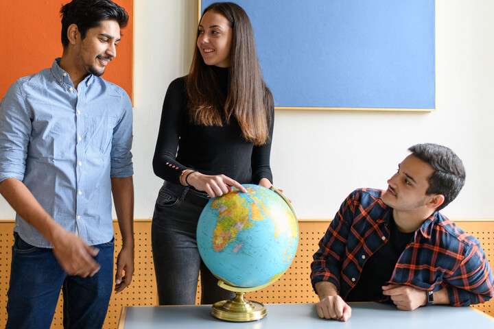 Drei Studierende um eine Weltkugel. Eine Studentin zeigt auf einen Punkt der Weltkugel.