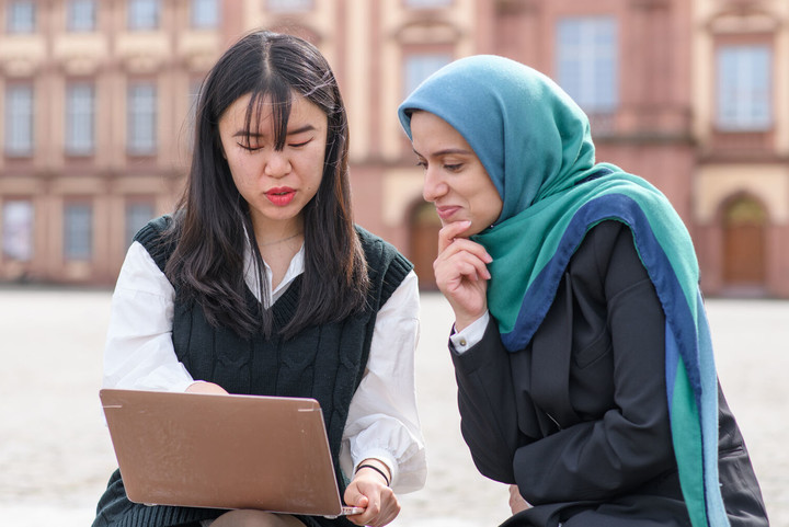 Zwei internationale Studentinnen sitzen auf dem Ehrenhof vor dem Schloss und blicken auf einen Laptopbildschirm