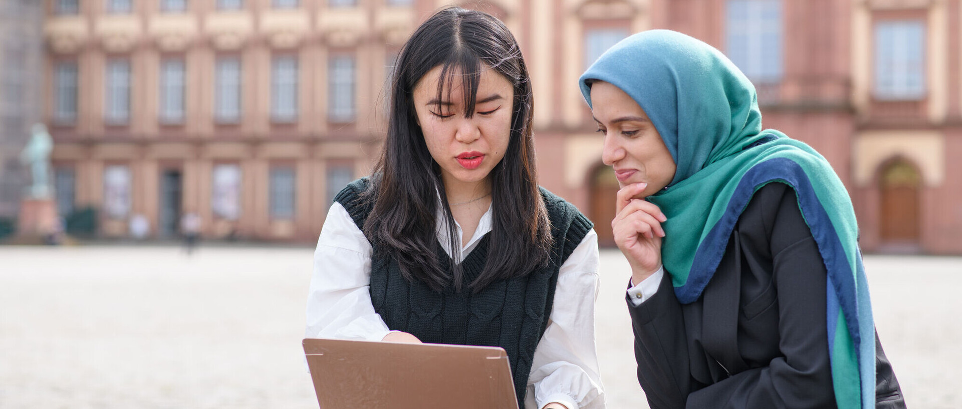 Zwei internationale Studentinnen sitzen auf dem Ehrenhof vor dem Schloss und blicken auf einen Laptopbildschirm