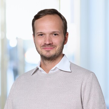 Benedikt Kastner hat kurze braune Haare und einen kurzen Bart. Er trägt ein beigefarbenes Sweatshirt und ein weißes Hemd.